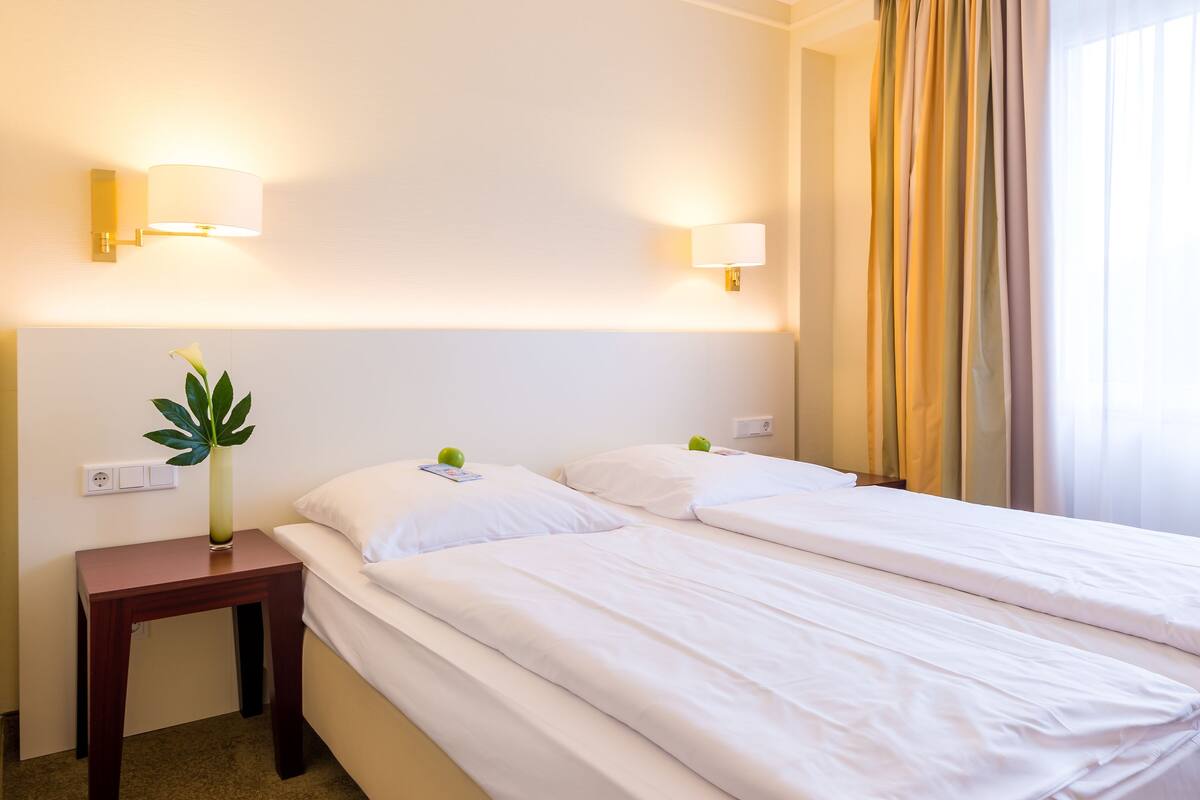 A hotel room with two beds, a nightstand, a lamp, and a window with curtains.