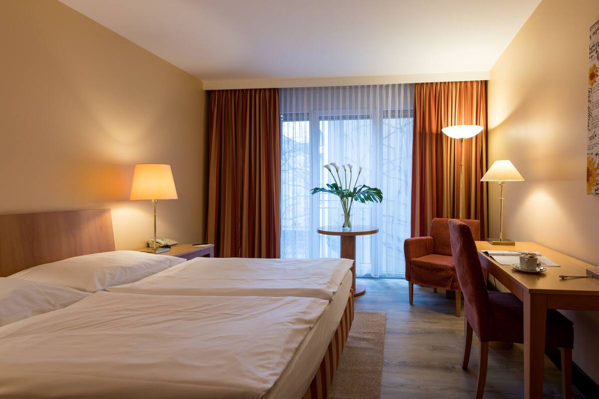 A hotel room with two beds, a desk, a chair, a lamp, and a window with curtains.