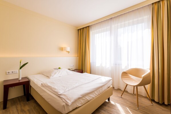 Una habitación de hotel con cama, mesita de noche, lámpara, silla y ventana con cortinas.