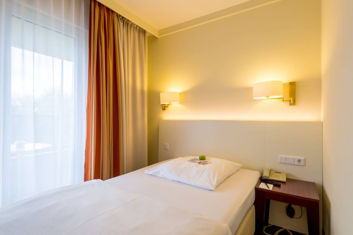 A hotel room with a bed, curtains, wall-mounted lamps, a bedside table with a telephone, and a window with sheer curtains.