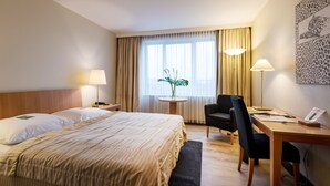 A hotel room with a large bed, a desk, two chairs, a small table with a plant, and a painting on the wall.