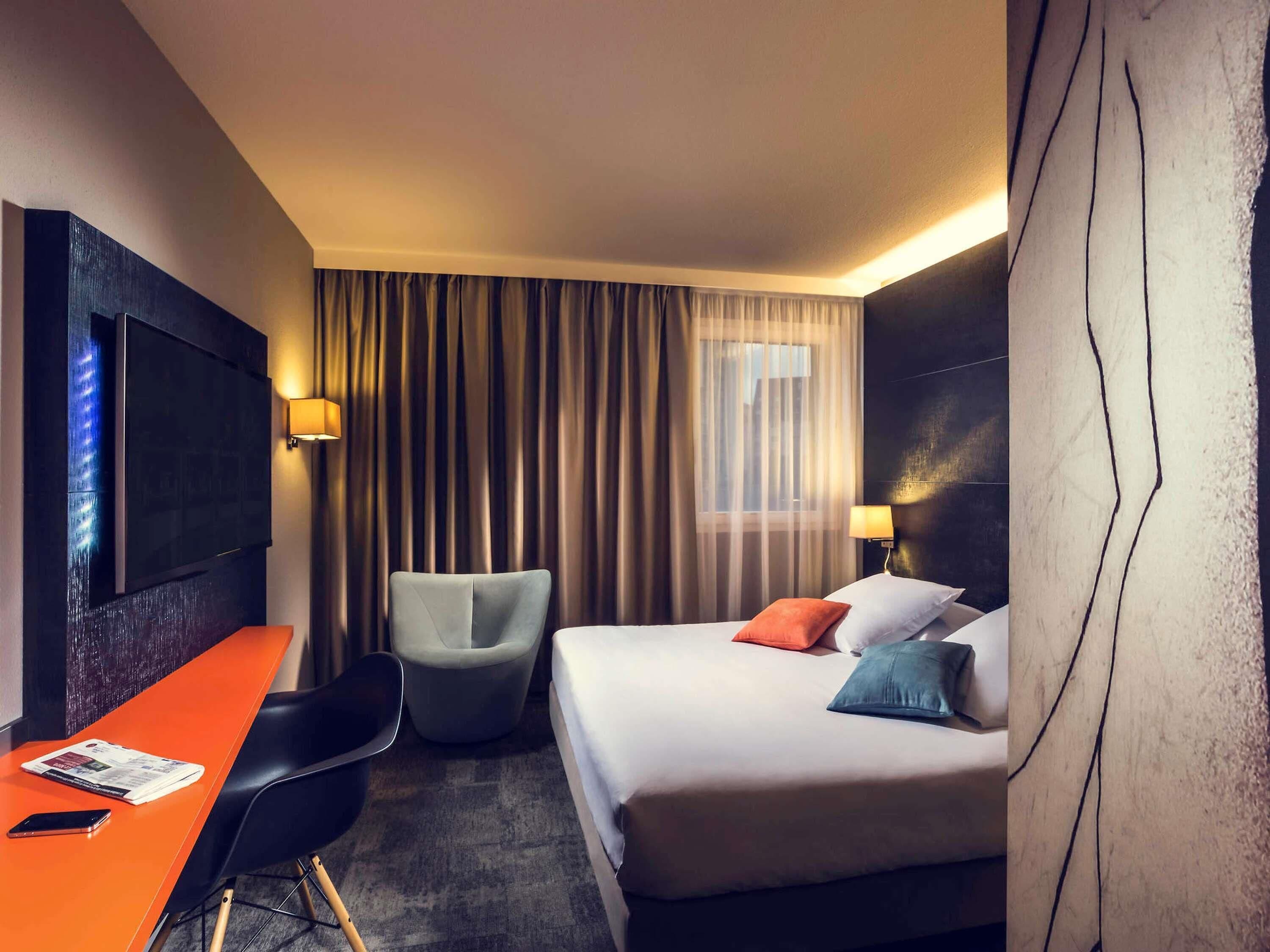 Una habitación de hotel con cama, escritorio, silla, televisión y ventana con cortinas.