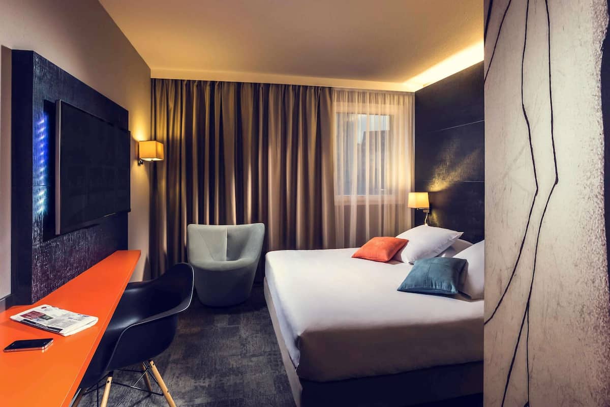 Una habitación de hotel con cama, escritorio, silla, televisión y ventana con cortinas.