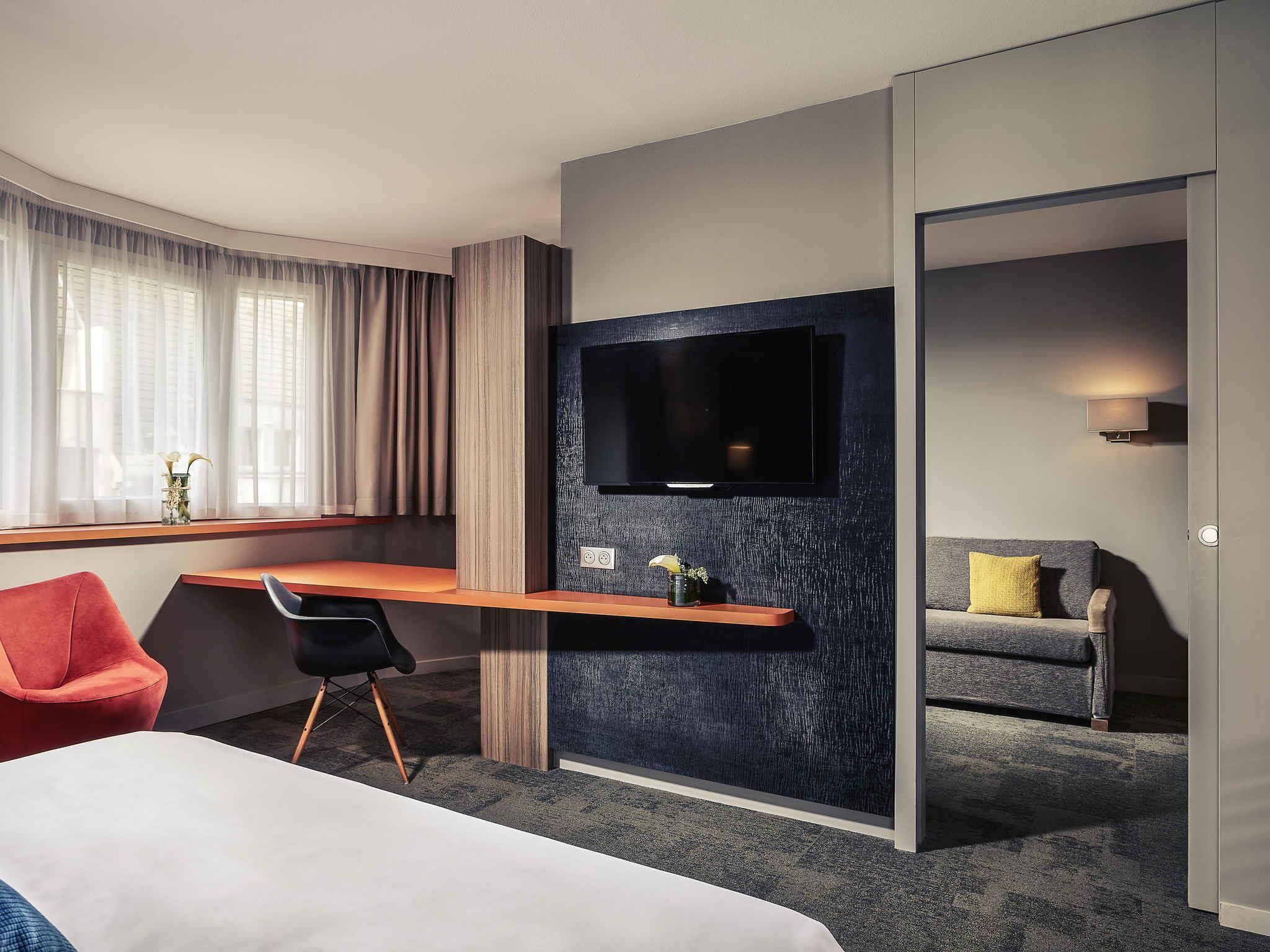 Habitación de hotel con cama, escritorio con televisión de pantalla plana, una silla roja, sofá gris y ventana con cortinas.