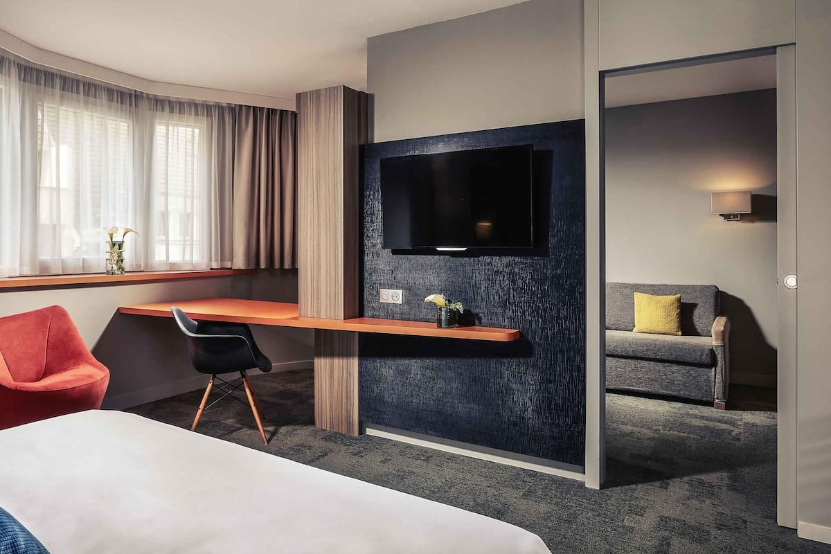 Habitación de hotel con cama, escritorio con televisión de pantalla plana, una silla roja, sofá gris y ventana con cortinas.