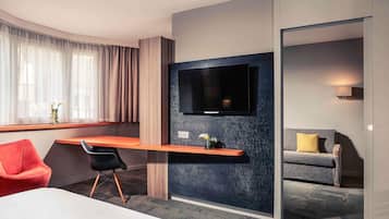 Una habitación de hotel moderna con una cama, un escritorio con silla, un televisor montado en una pared texturizada, un sofá y una ventana con cortinas.