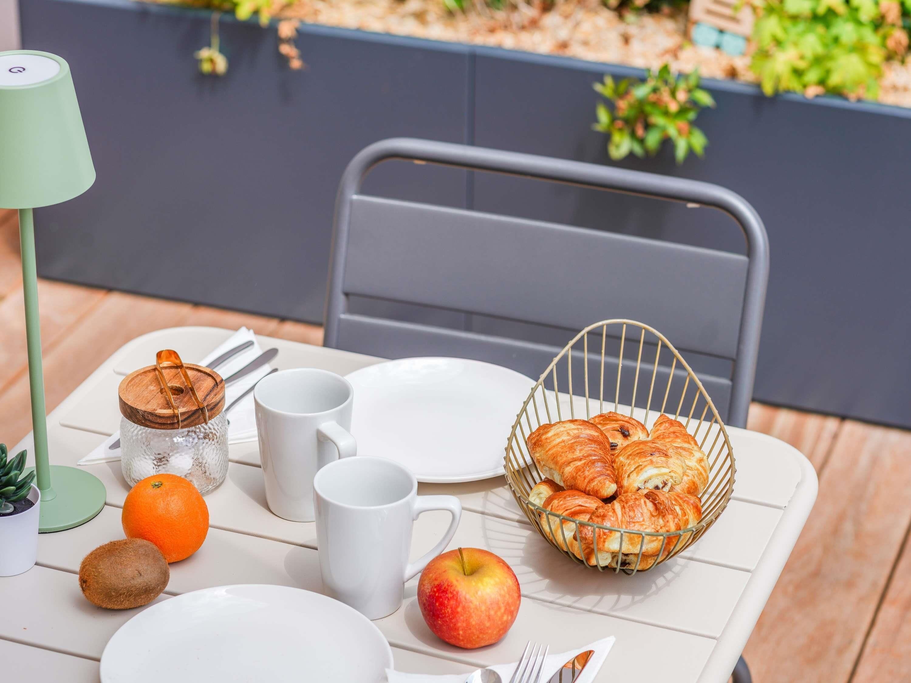 Una mesa dispuesta con platos, tazas, una cesta de cruasanes, una manzana, una naranja y un tarro con tapa.