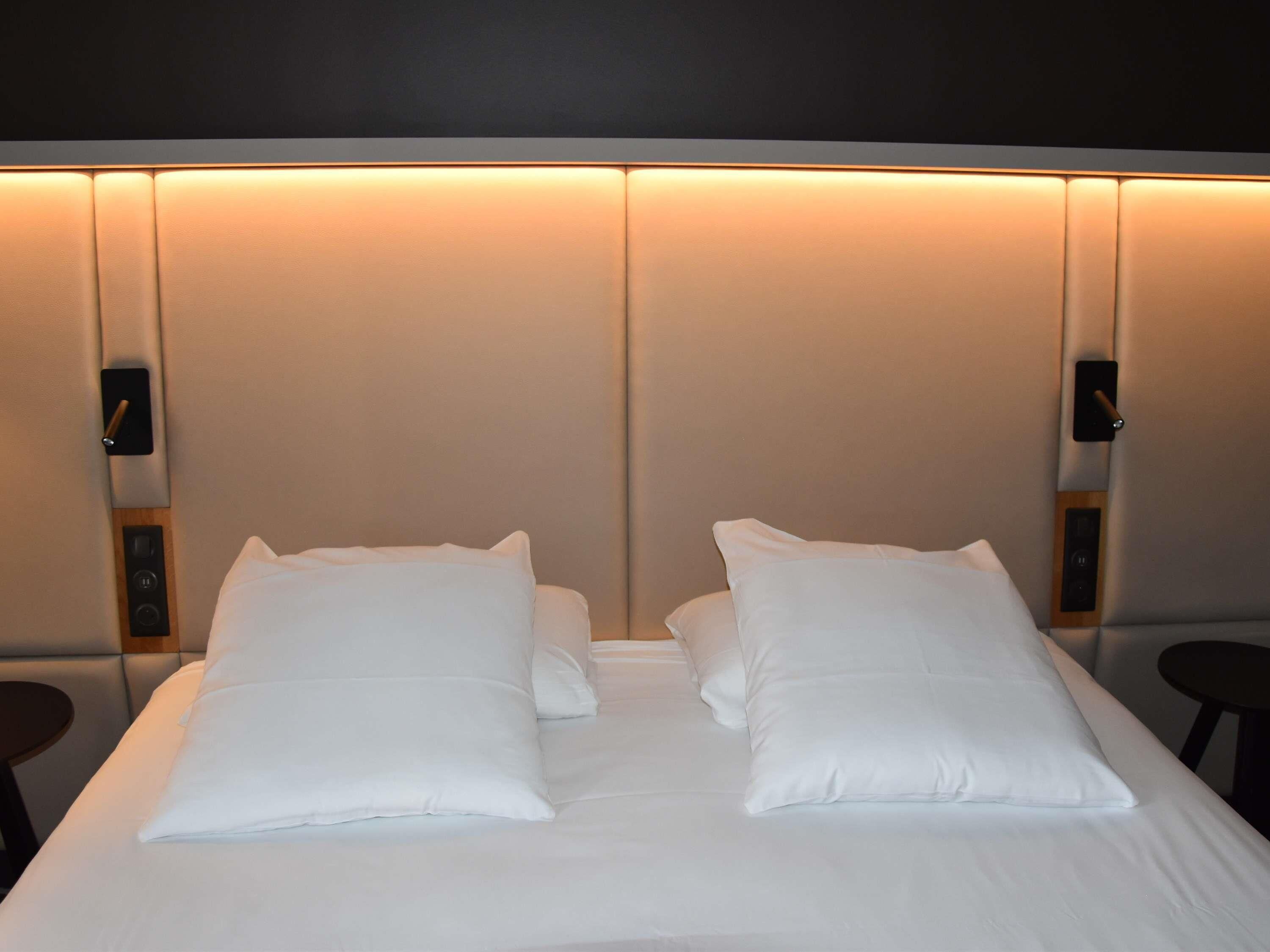 Una cama impecablemente hecha con sábanas y almohadas blancas, un cabecero con iluminación integrada y dos mesitas de noche con lámparas negras.