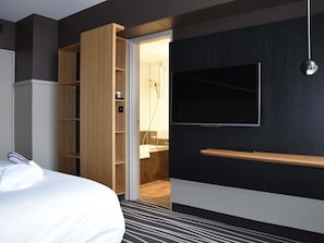 Habitación de hotel moderna con cama, armario, televisión y baño visible a través de una puerta abierta.
