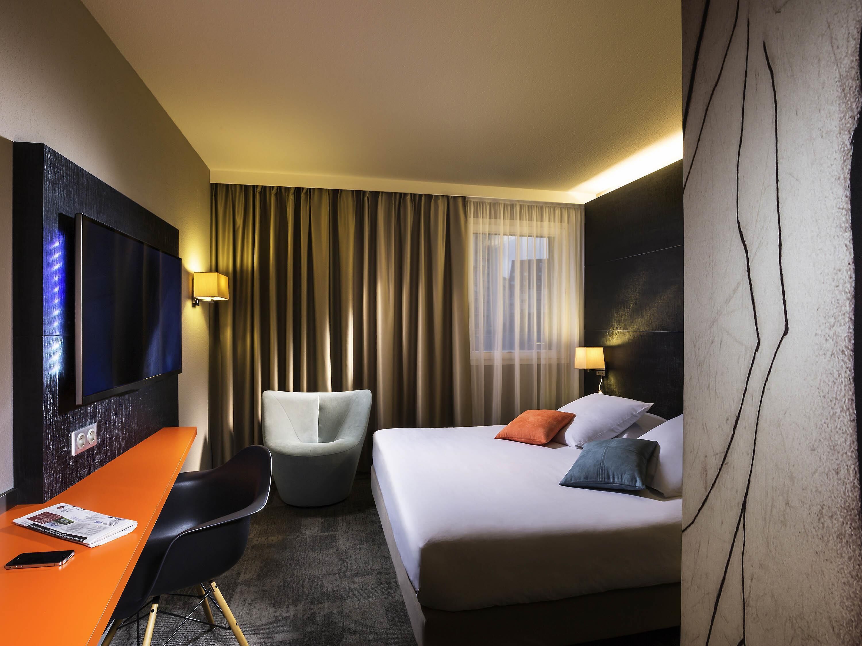 Une chambre d’hôtel avec un lit, un bureau, une chaise, une télévision et une fenêtre avec des rideaux.