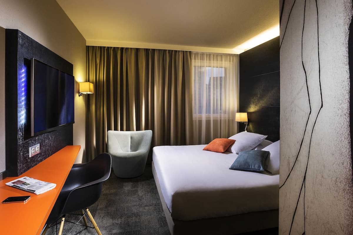 Une chambre dâhĂŽtel avec un lit, un bureau, une chaise, une tĂ©lĂ©vision et une fenĂȘtre avec des rideaux.