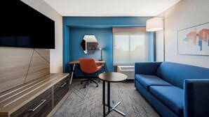 A modern hotel room with a blue accent wall, a flat-screen TV mounted on the wall, a desk with a chair, a round table, a sofa, and a window with a view. - Holiday Inn Express Hampton - Coliseum Central by IHG (Hampton)