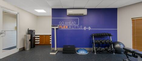 Une salle de sport avec un mur bleu, un climatiseur fixé au mur, un porte-haltères, une plateforme de step et un banc.