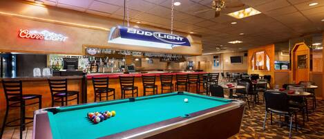 A pool table with a neon sign for Bud Light, a bar area with stools, and a seating area with tables and chairs.