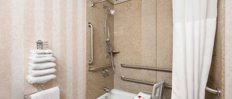 Ein Badezimmer mit Dusche, Haltegriffen, einer rollstuhlgerechten Sitzbank und gestapelten Handtüchern.