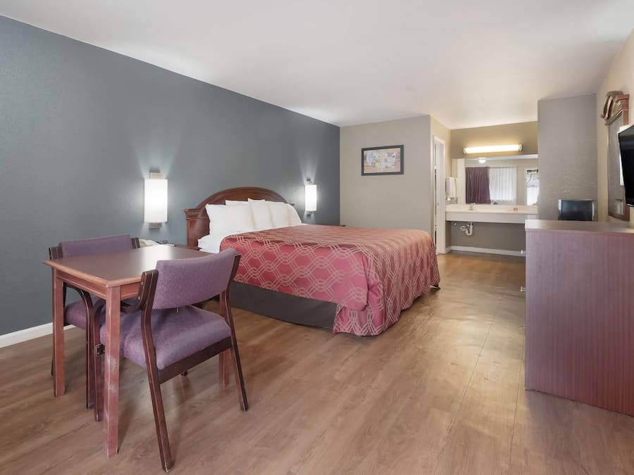 Quarto de hotel com uma cama, uma escrivaninha com cadeira, uma televisão e um banheiro visível ao fundo.