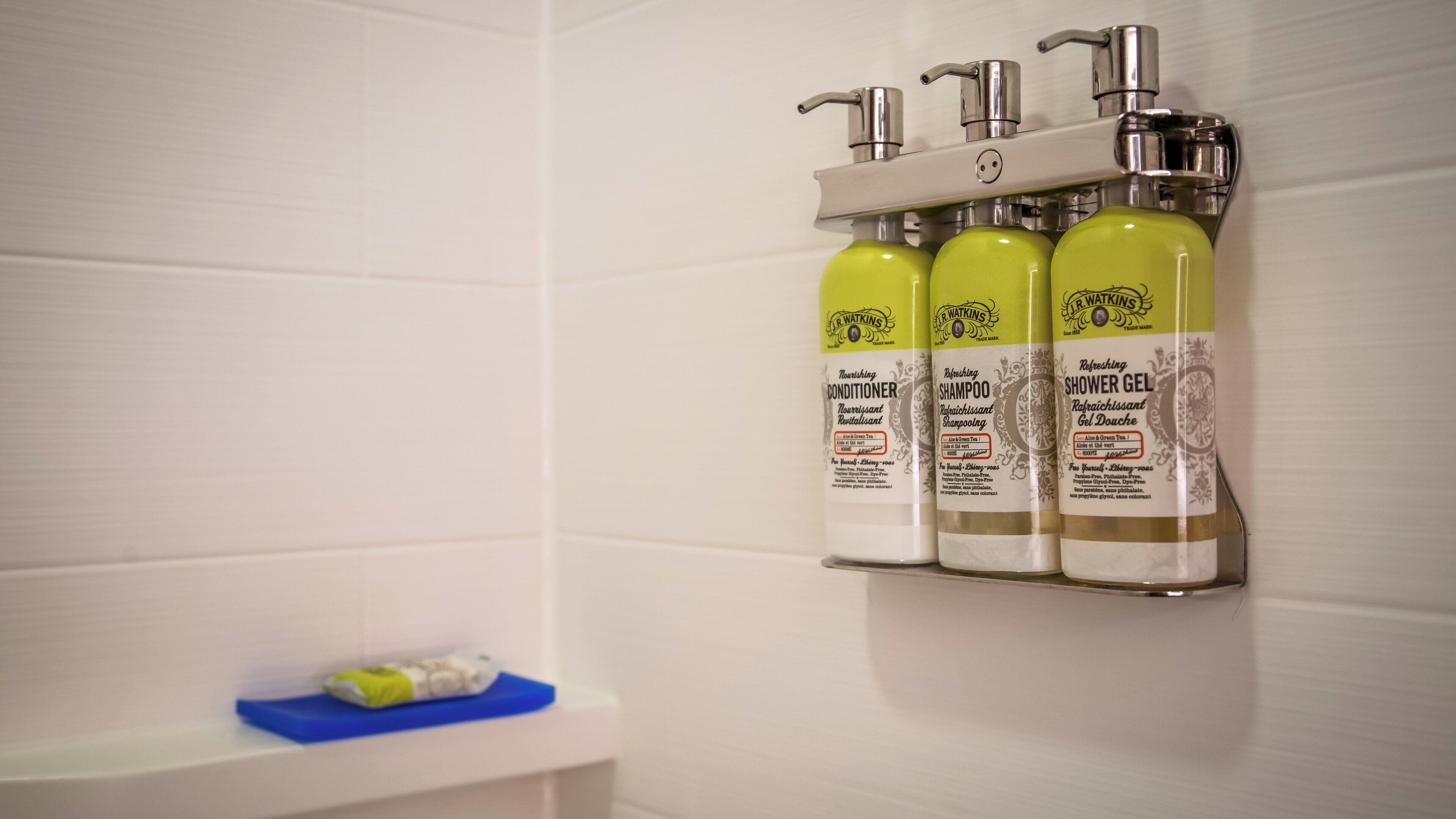 A bathroom with a white tiled wall, a shelf with three bottles of R. Watkins products, and a blue soap dish.