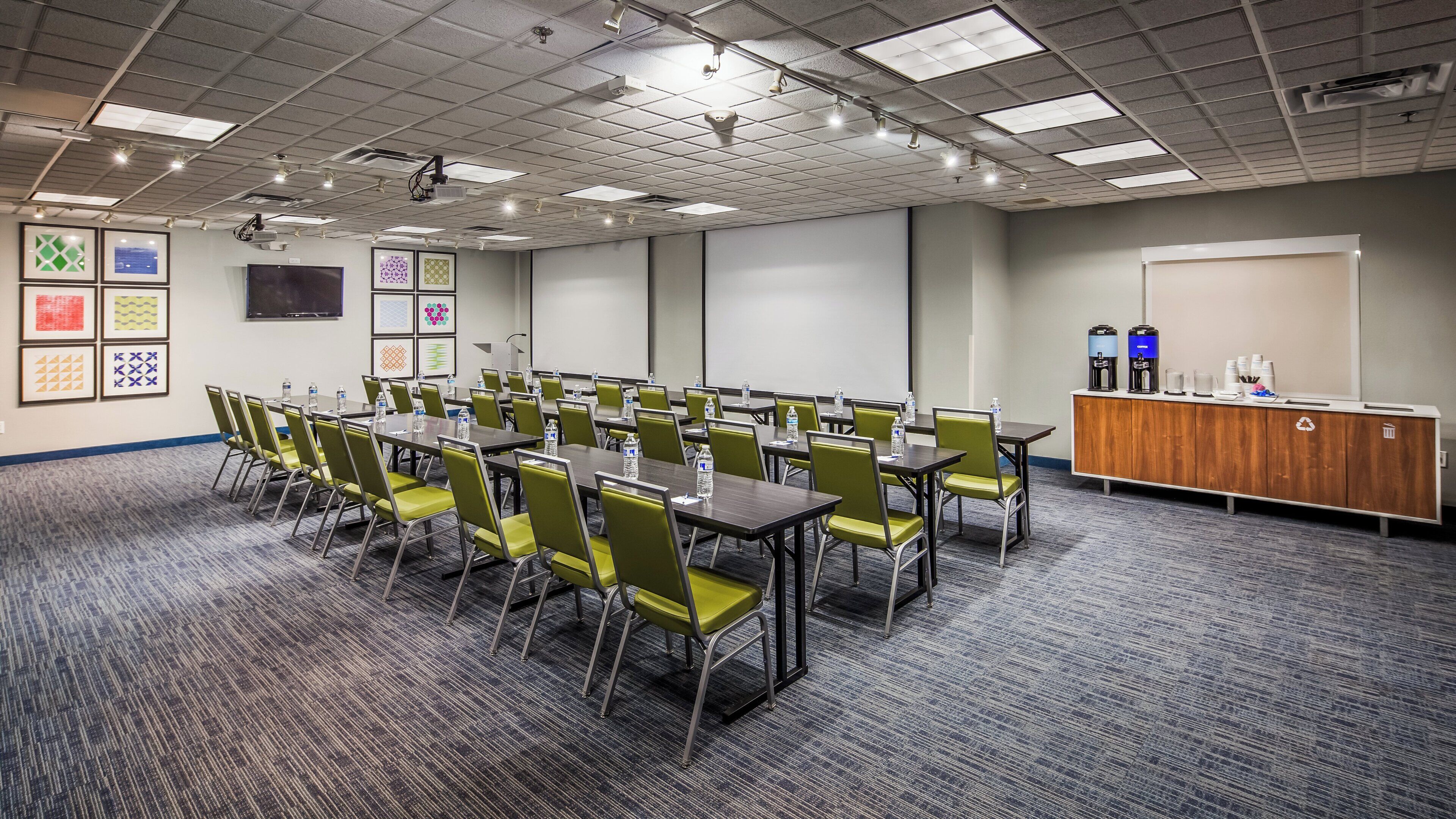 A conference room with tables and chairs, a projector, a screen, and a podium with water bottles and coffee.