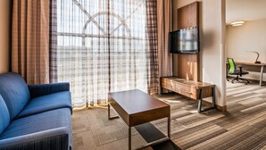 Une chambre d’hôtel avec un canapé, un téléviseur fixé à un meuble en bois, un bureau avec une chaise et une moquette à motifs.