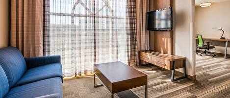 Quarto de hotel com sofá, televisão fixada em um móvel de madeira, escrivaninha com cadeira e carpete estampado.