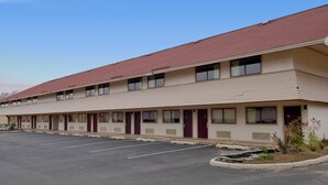A multi-story motel with a red roof, a parking lot, and a small landscaped area with plants.