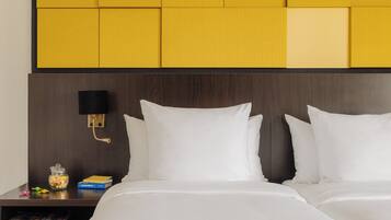 A hotel room with a wooden headboard, two white pillows, a bedside table with a lamp, a book, and a jar of candies.