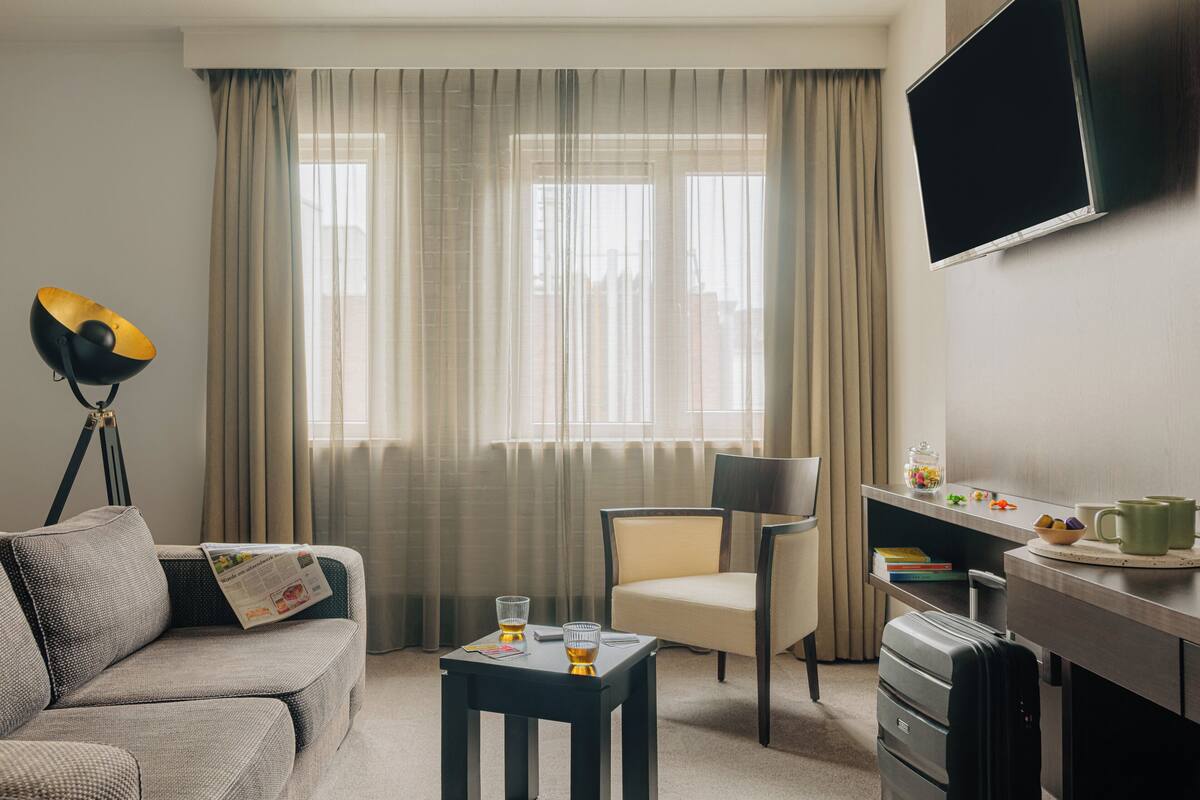Habitación de hotel con sofá, una mesita con vasos, una silla, escritorio con televisor de pantalla plana y ventana con cortinas.