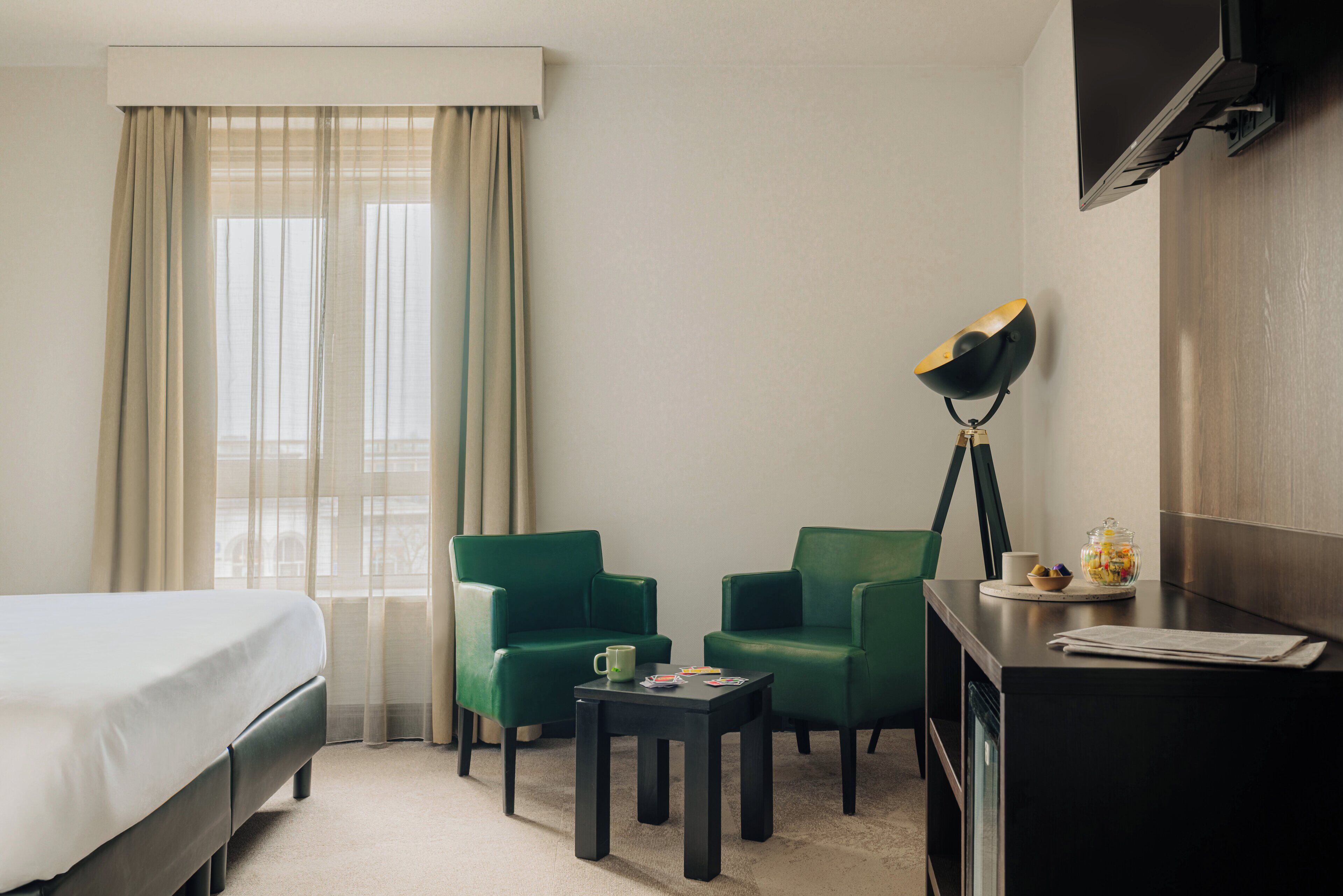 A hotel room with a bed, two green armchairs, a small table, a lamp, and a TV mounted on the wall.