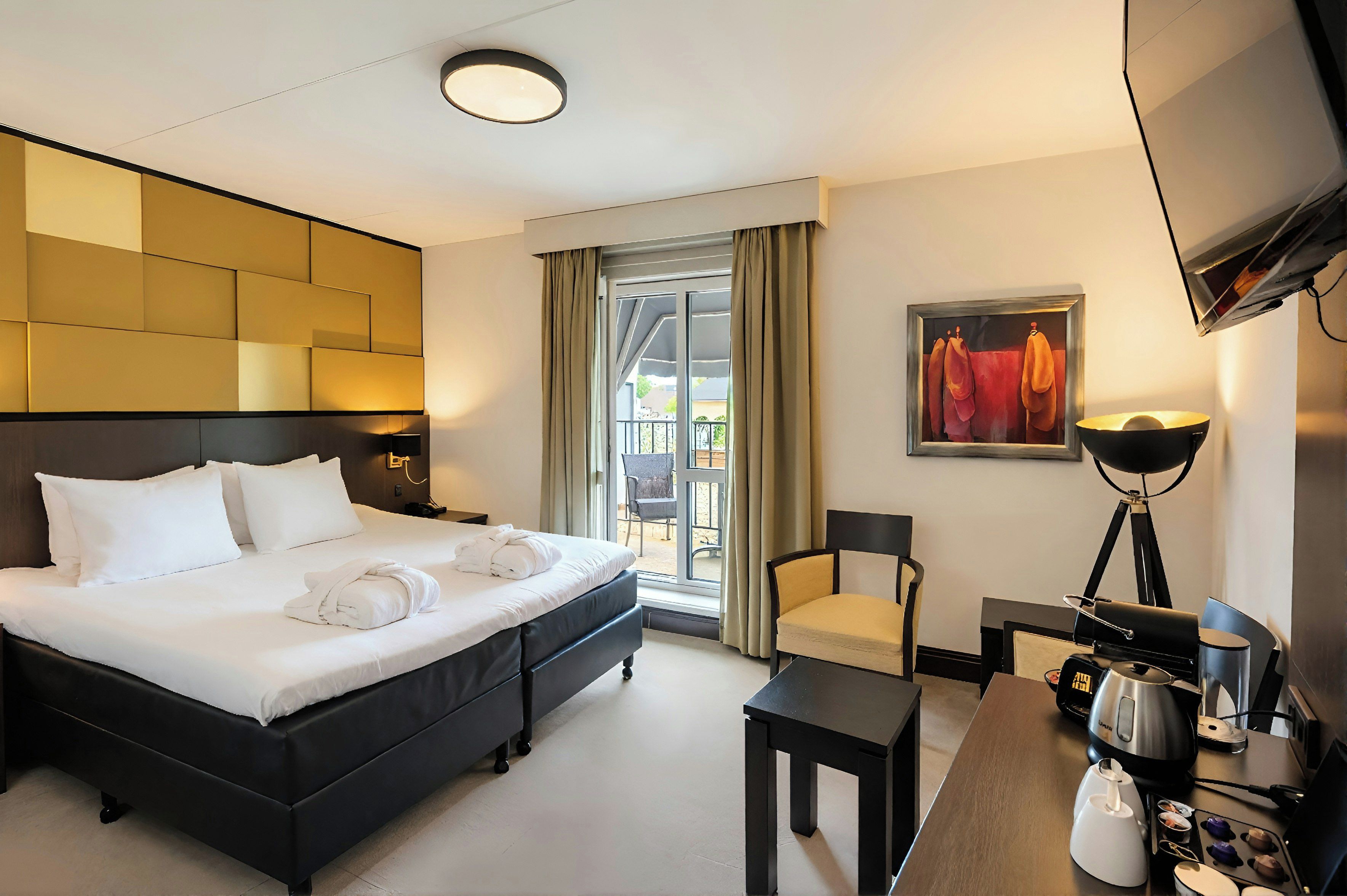 Une chambre d’hôtel avec un grand lit, un bureau avec une bouilloire, une chaise et un tableau au mur.