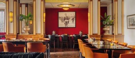 Un restaurante con paneles de madera, paredes rojas y sillas naranjas. Hay mesas con sillas, un cuadro grande en la pared y plantas en macetas.