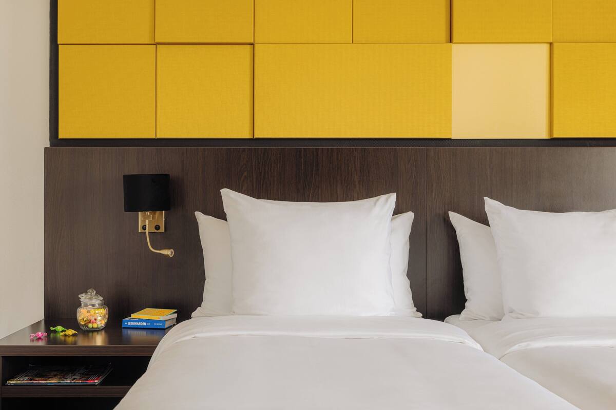 Una habitación de hotel con cabecera de madera, dos almohadas blancas, una mesita de noche con lámpara, un libro y un tarro de caramelos.