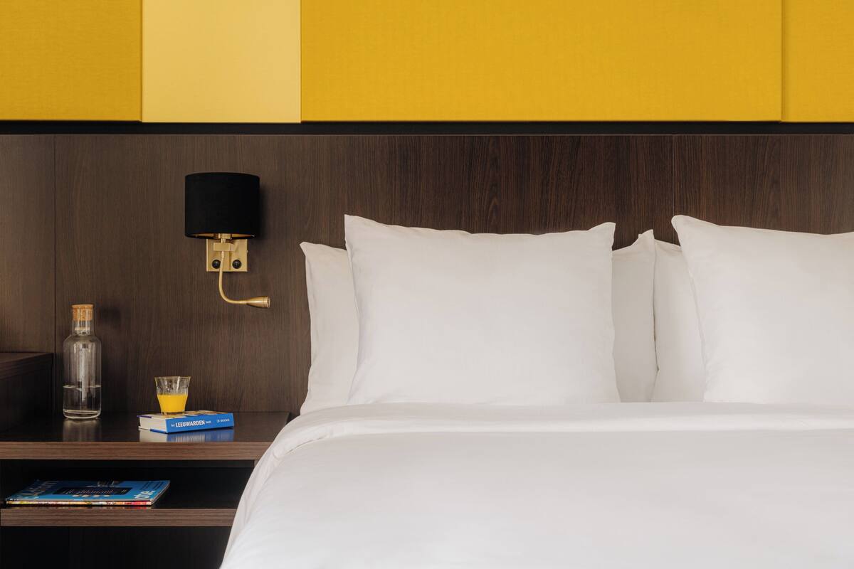 Habitación de hotel con cabecera de madera, ropa de cama blanca, mesita de noche con un vaso de jugo de naranja, una botella y un libro.