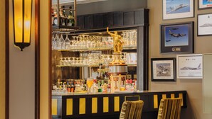 A bar area with a counter, glasses, bottles, and a statue. There are chairs and framed pictures on the wall.