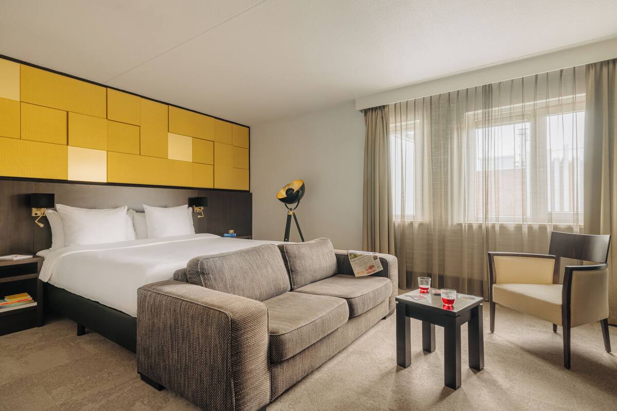 Habitación de hotel con cama, sofá, una mesita, una silla, lámpara y ventana con cortinas.