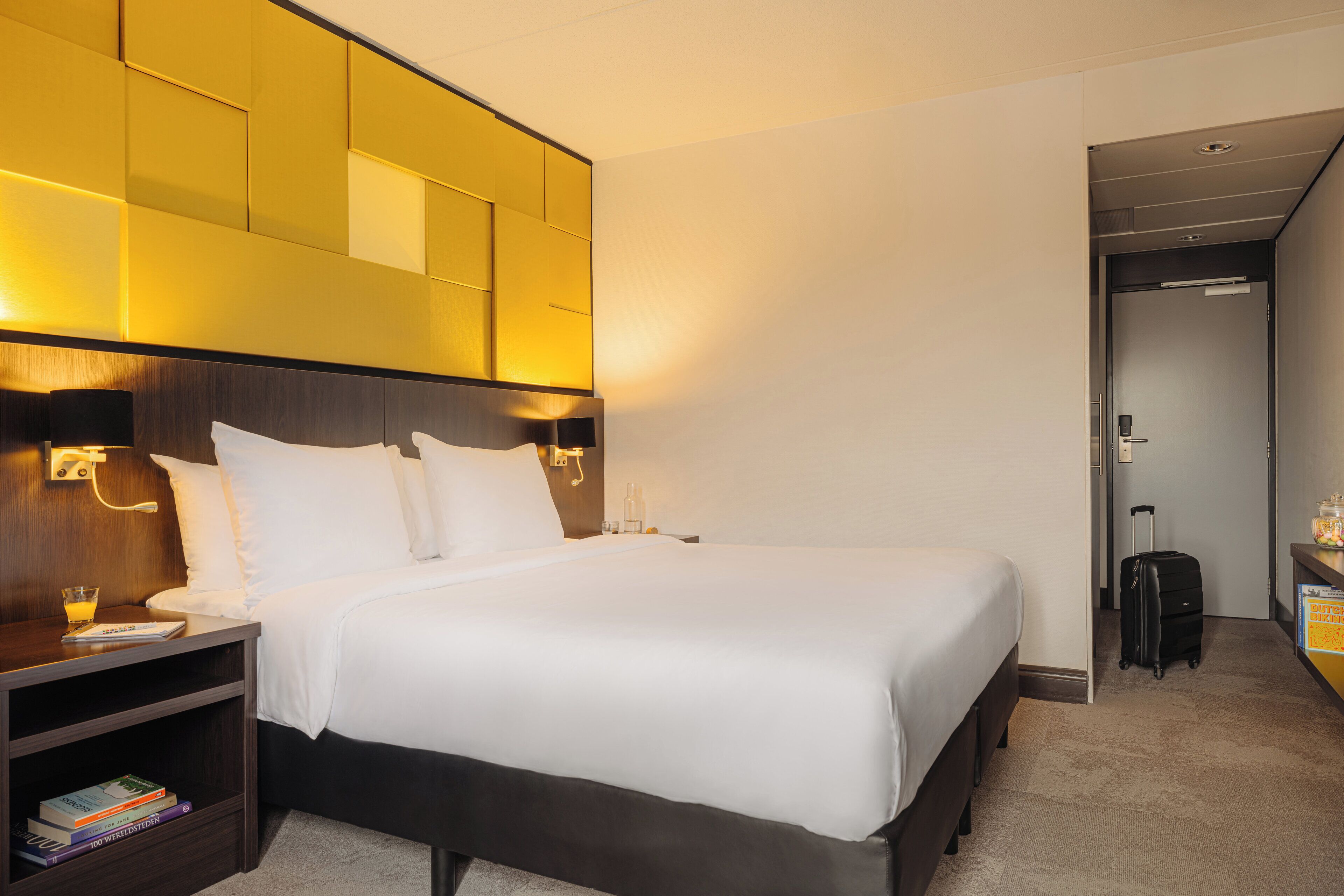 A hotel room with a large bed, bedside tables, a nightstand, a wall-mounted light, and a suitcase in the corner.