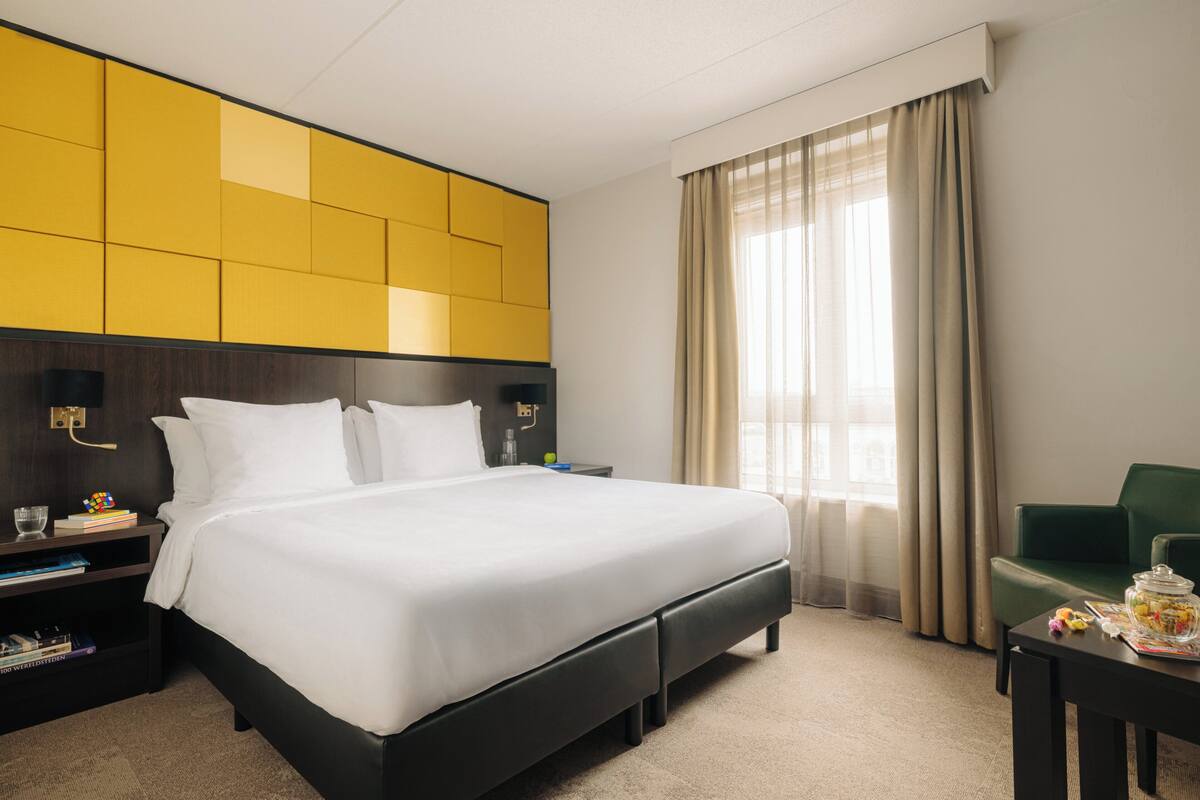 Una habitación de hotel con una cama grande, una mesita de noche, un sillón verde y una ventana con cortinas.