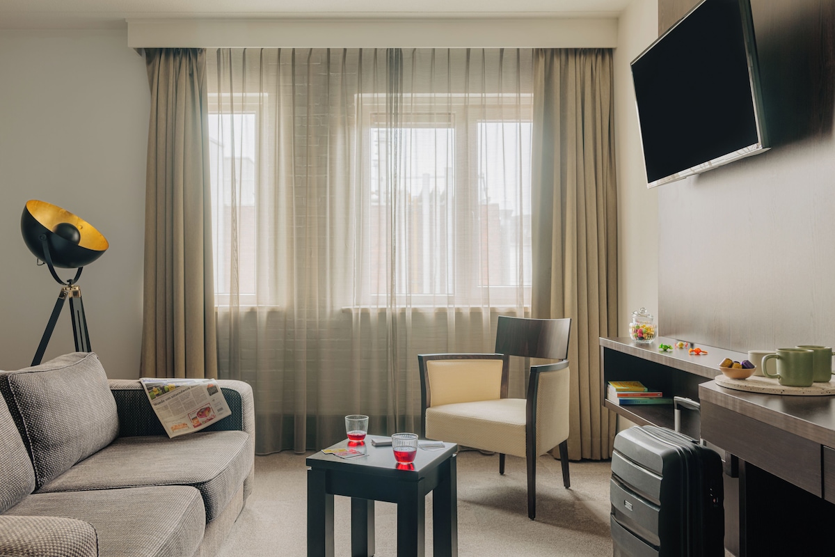 Habitación de hotel con sofá, una mesita con bebidas, una silla, escritorio con televisor y ventana con cortinas.