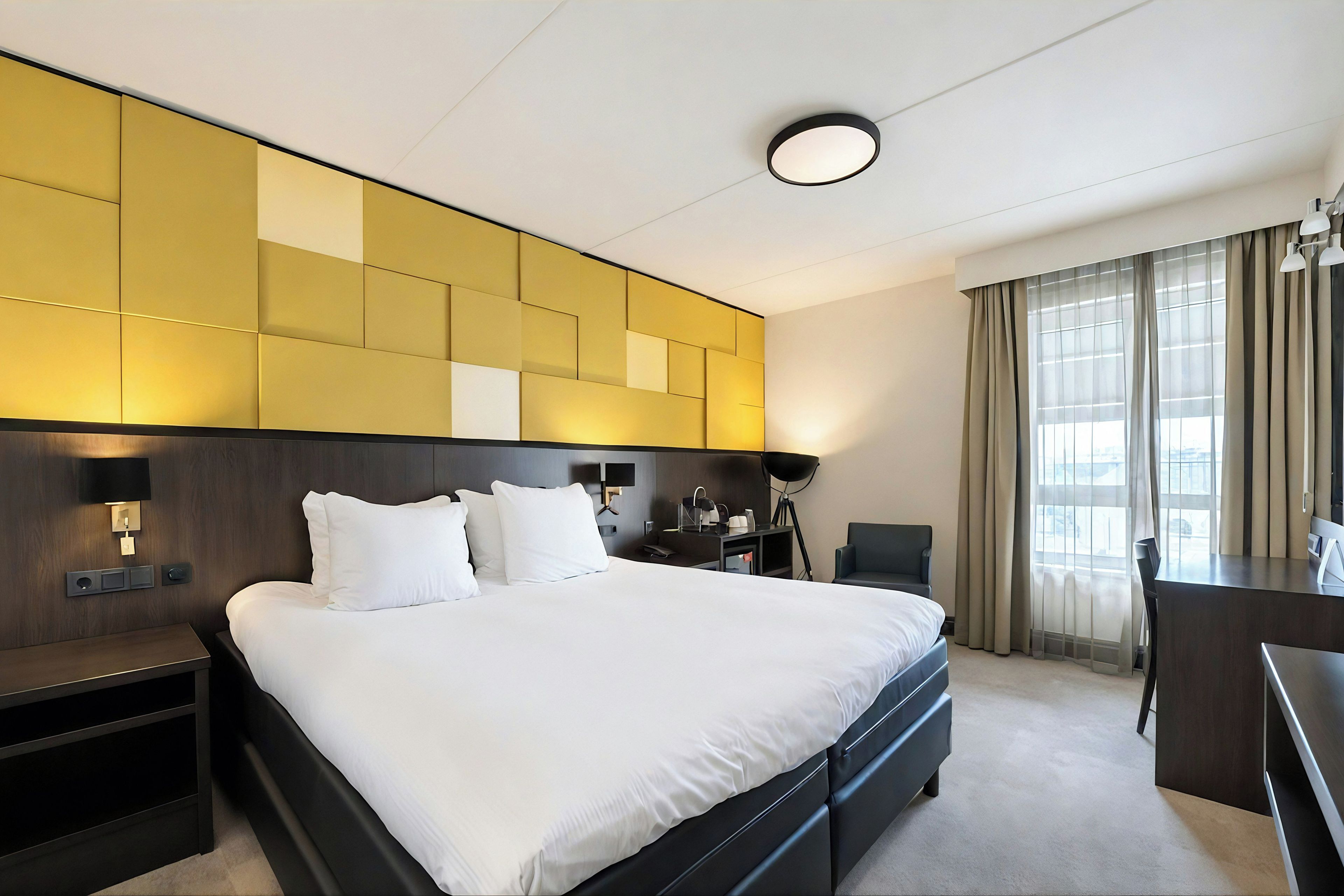 Una habitación de hotel con una cama grande, mesitas de noche, un escritorio, una silla y una ventana con cortinas.