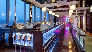 Un bar moderne doté d’un long comptoir, de nombreux tabourets et offrant une vue sur la ville grâce à de grandes fenêtres.