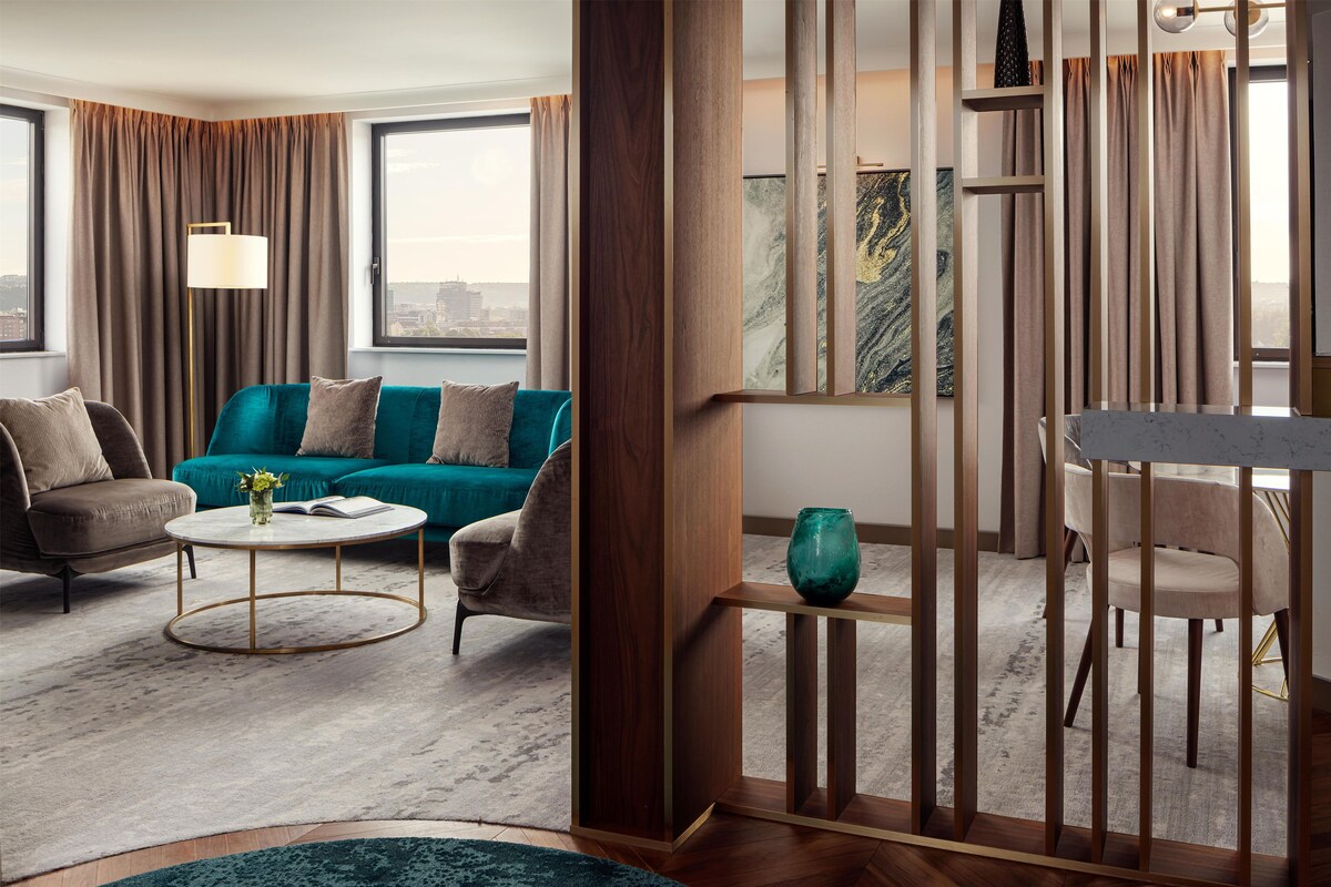 A modern living room with a teal sofa, a round coffee table, and a view of the city through large windows.