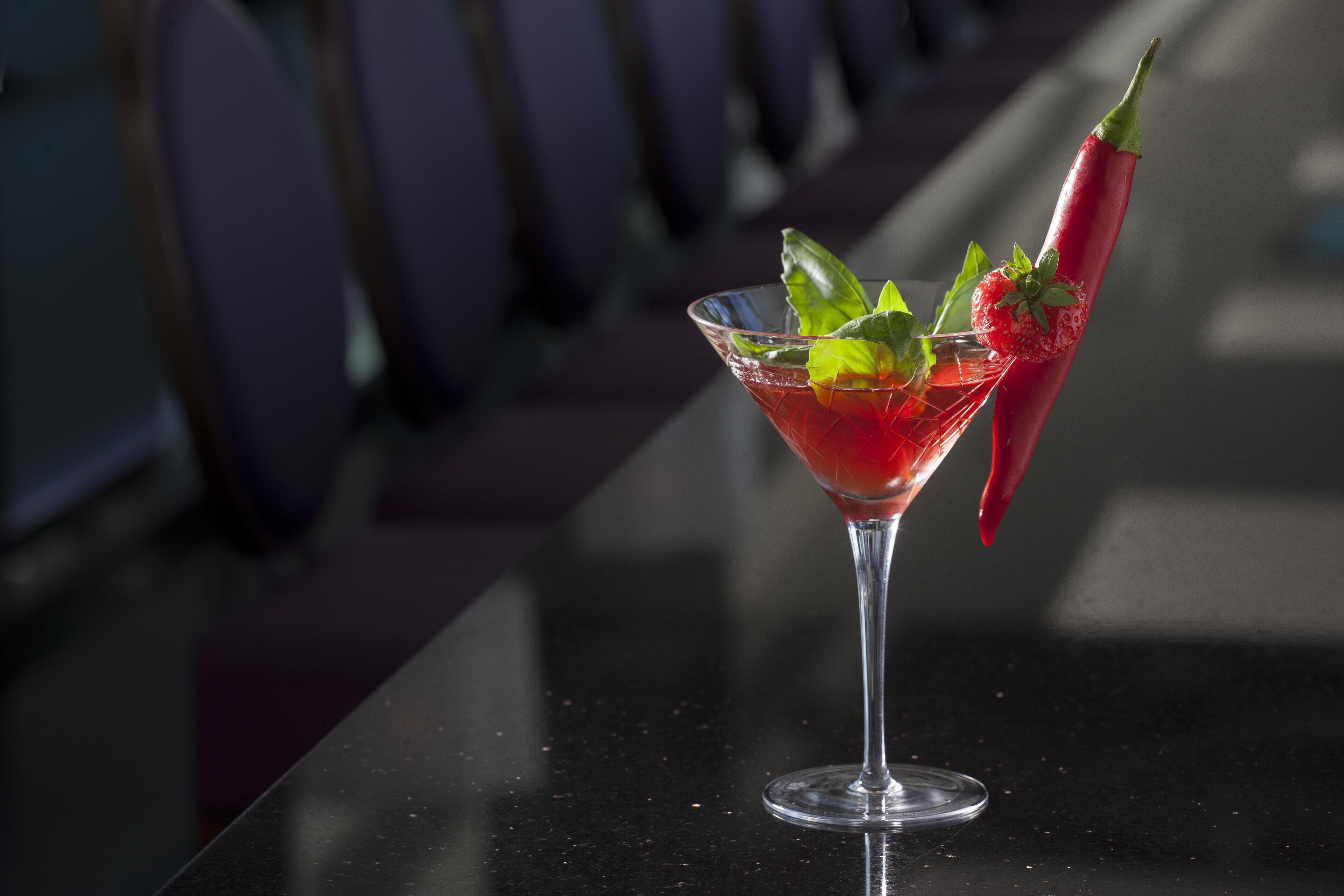 A martini glass with a red drink, garnished with a strawberry and a chili pepper, on a reflective black table.