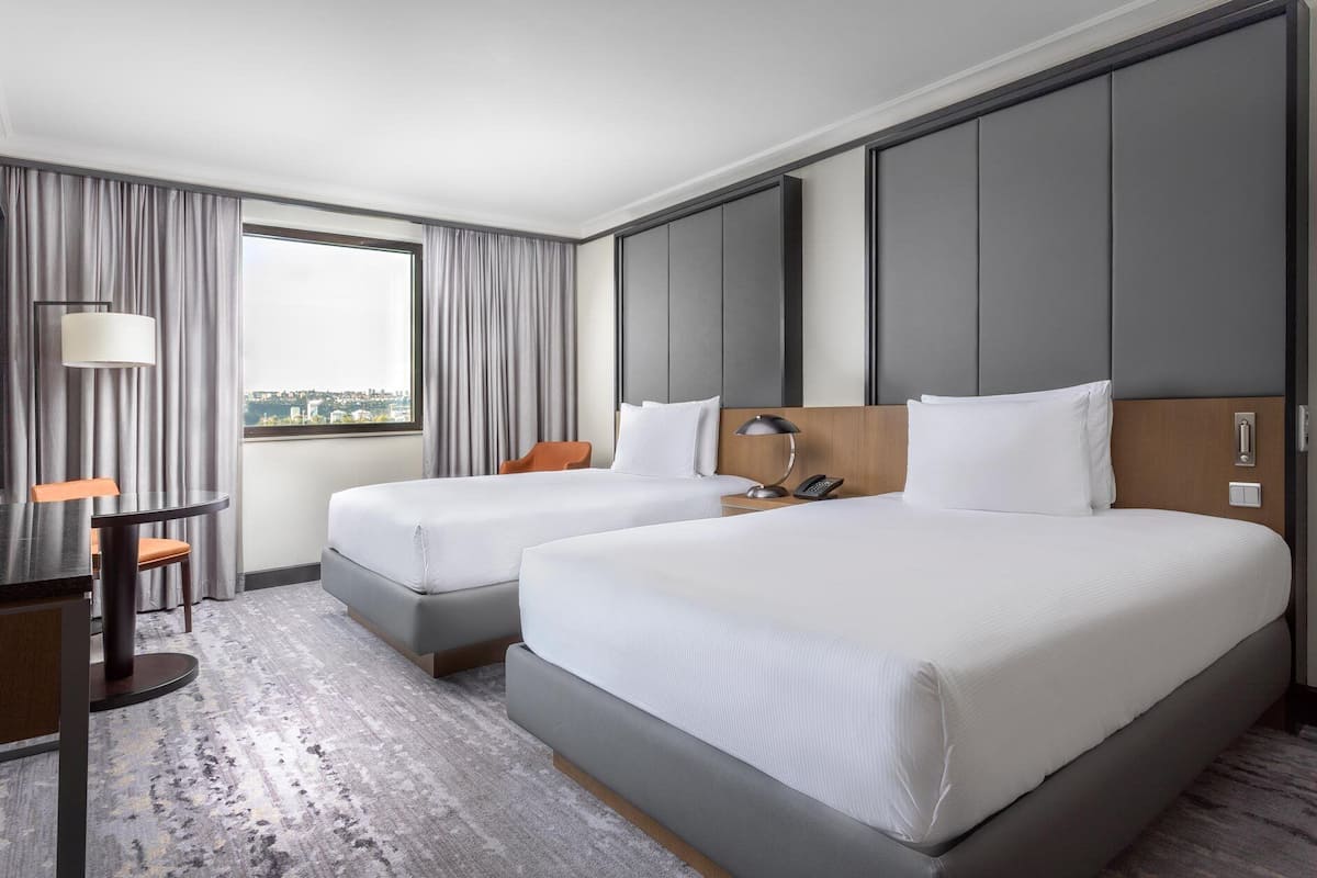 Ett hotellrum med två sängar, ett skrivbord, en stol, en lampa och utsikt över staden.