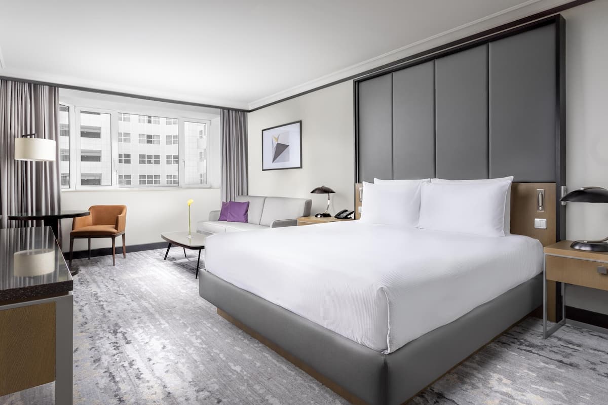 Ett modernt hotellrum med en stor säng, ett skrivbord, en stol, en lampa och ett fönster med utsikt  över staden.