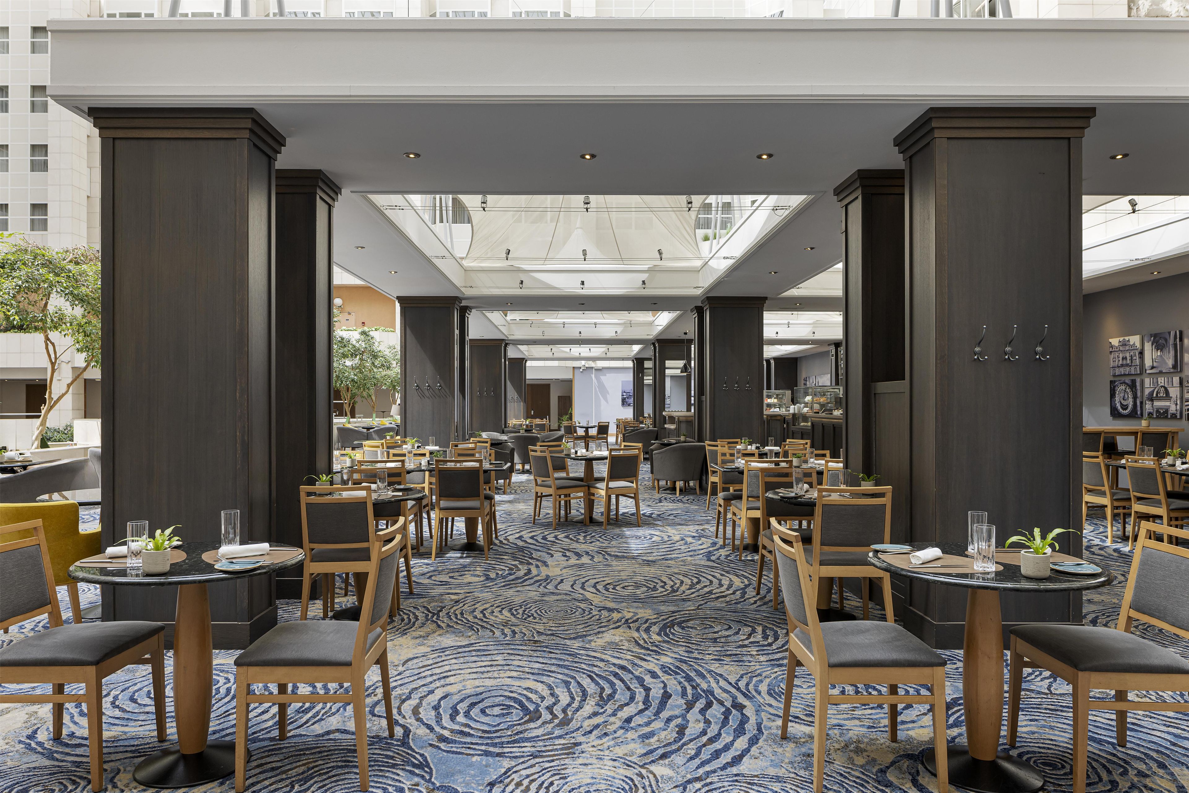 En rummelig hotelrestaurant med flere borde og stole, et mønstret tæppe og et højt loft med naturligt lys.