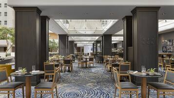 En rummelig hotelrestaurant med flere borde og stole, et mønstret tæppe og et højt loft med naturligt lys.