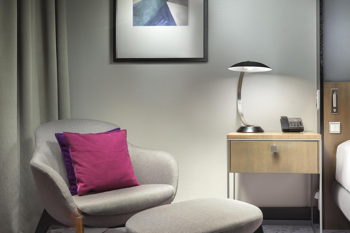 Ett modernt hotellrum med en grå fåtölj, ett litet sidobord med en lampa, en telefon och en säng.