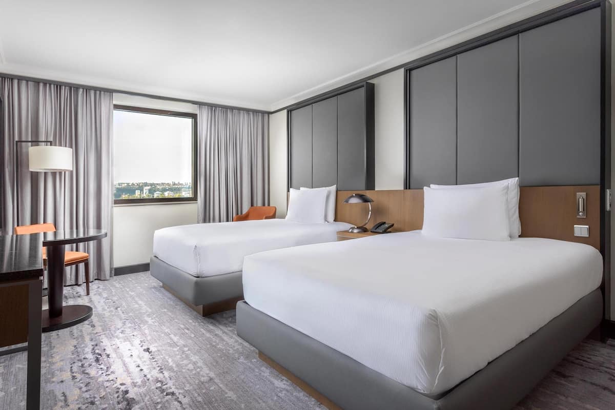 Ett hotellrum med två sängar, ett skrivbord, en stol, en lampa och utsikt över staden genom fönstret.