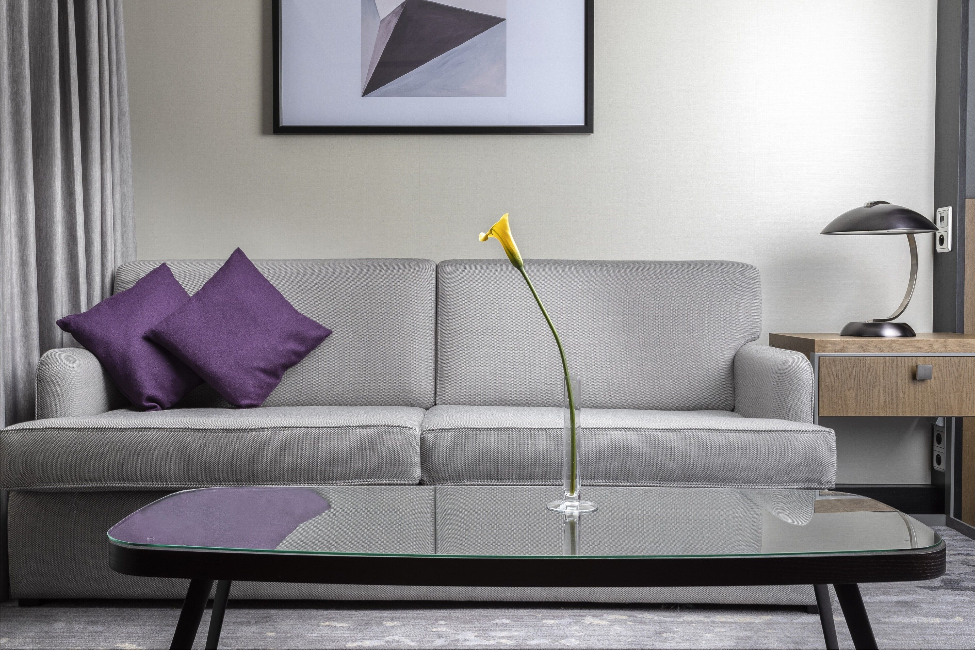 Un salon moderne avec un canapé gris, des coussins violets, une table basse en verre et une fleur jaune.