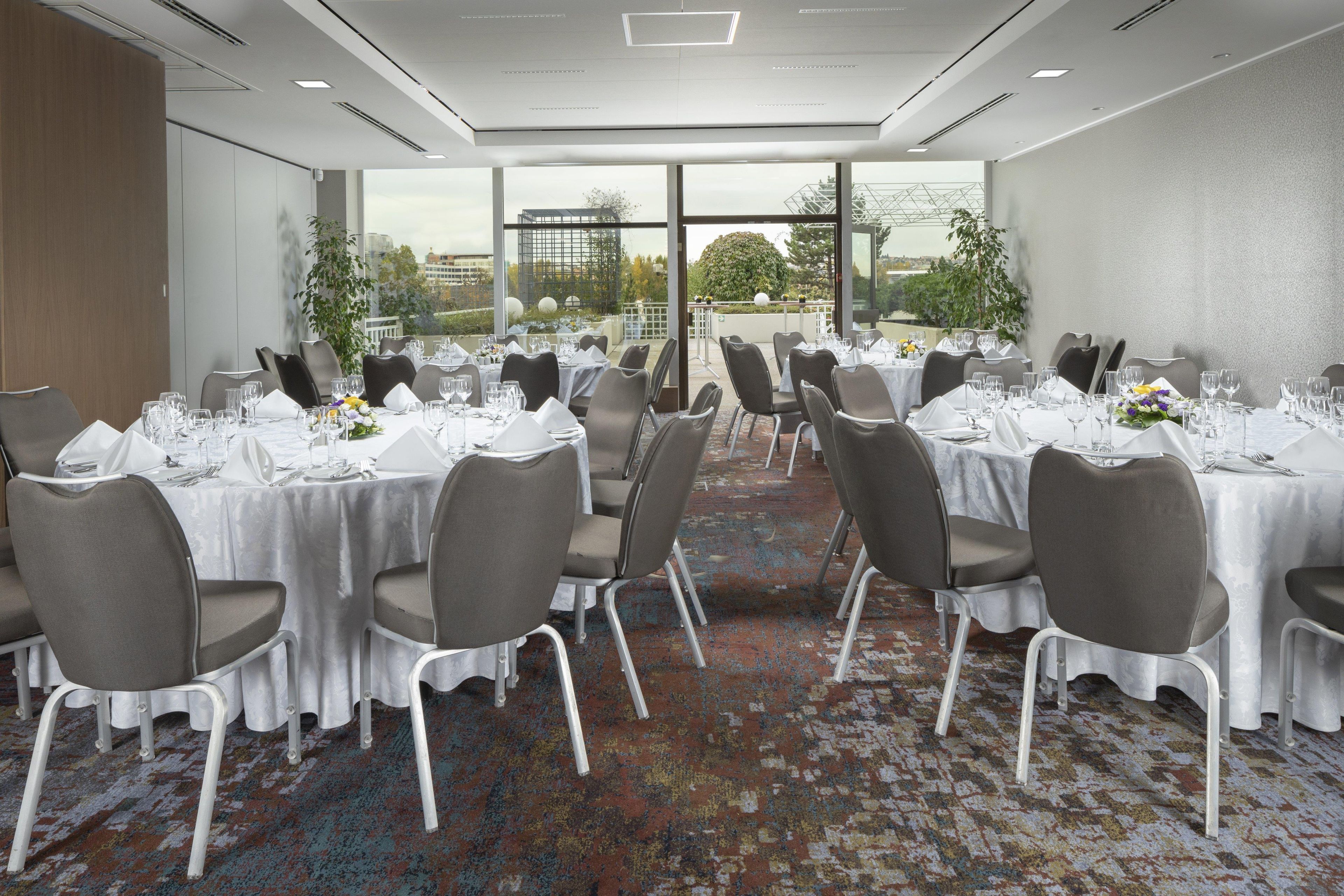 Sala de conferências com várias mesas preparadas para refeições, cadeiras e piso coberto por carpete.
