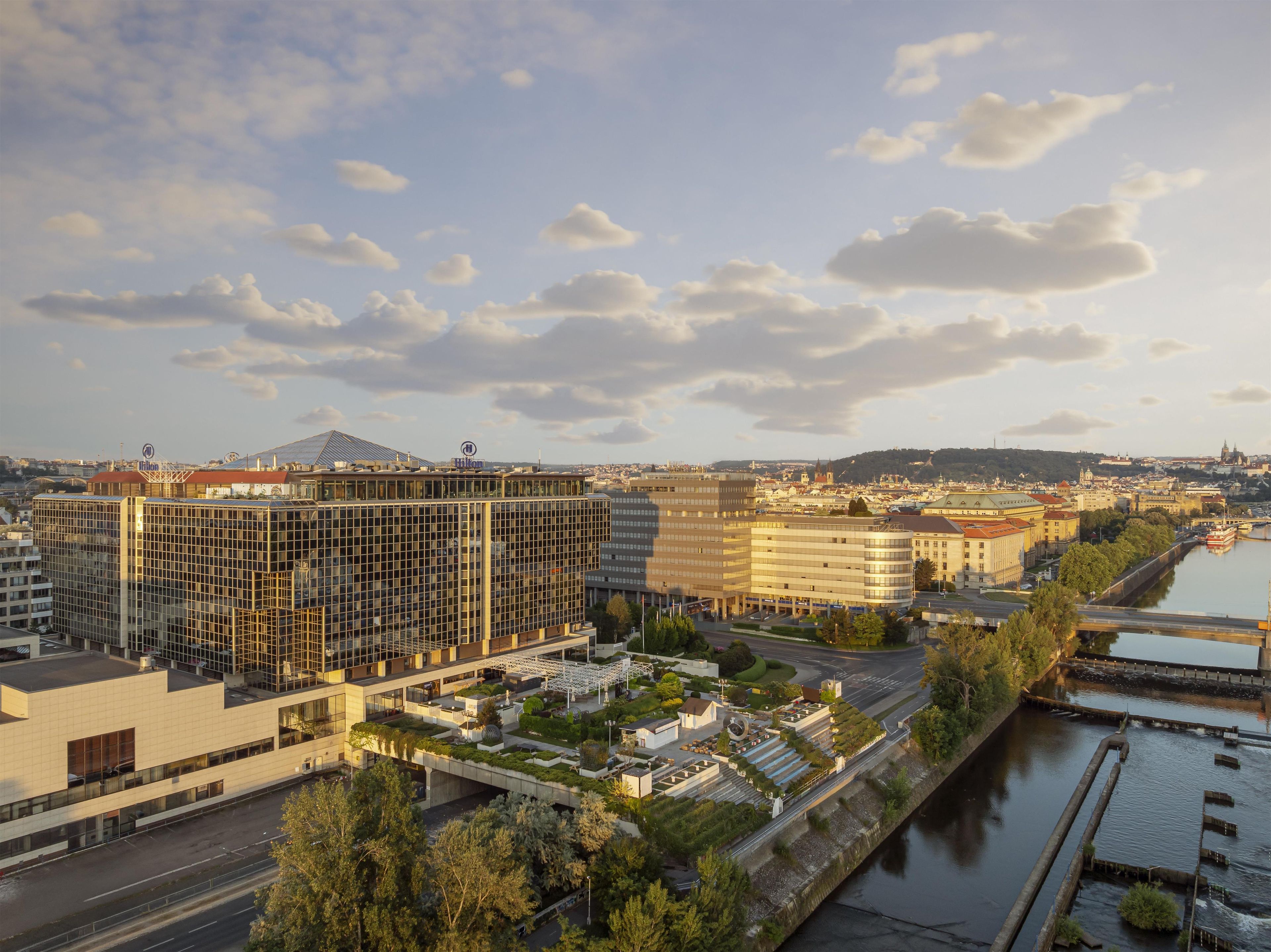 A modern hotel with a glass facade, located near a river with a bridge and a cityscape in the background.